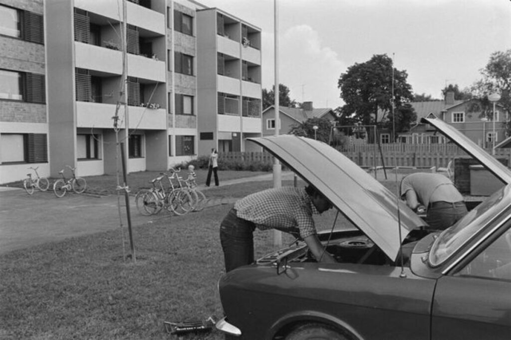 Mustavalkoinen valokuva autosta, jonka konepelti on auki ja mies korjaa sen alla moottoria.